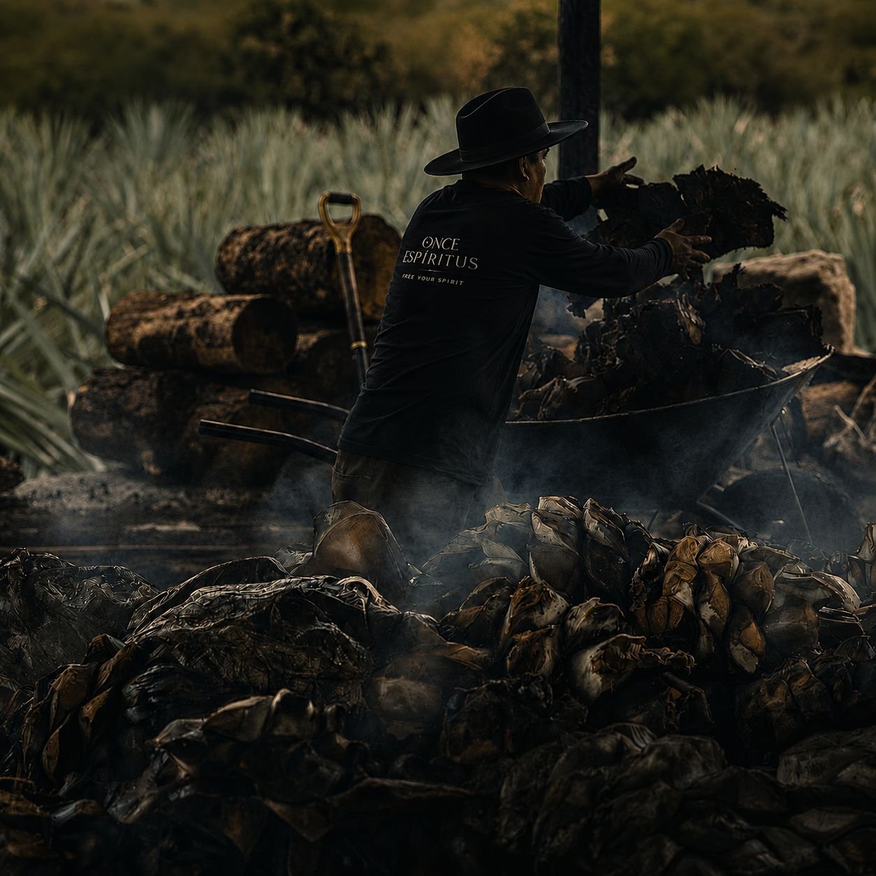 Un hombre con playera de Once Espíritus alimenta el horno de tierra entre el humo, con las piñas tatemadas en primer plano
